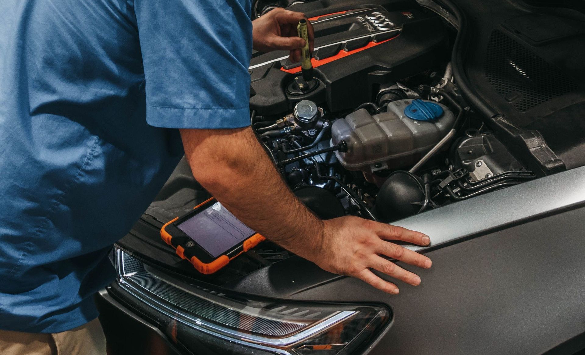 Mechanic in a workshop examining a car's engine under an open hood.
